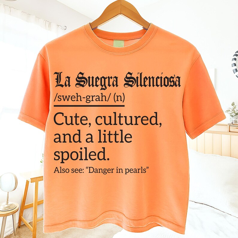 Bright orange t-shirt on a hanger with black text reading “La Suegra Silenciosa /sweh-grah/ (n) Cute, cultured, and a little spoiled. Also see: ‘Danger in pearls,’” displayed in a bedroom.