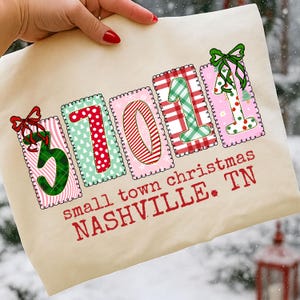 May include: A beige tote bag with the text "small town christmas NASHVILLE. TN". The bag features the numbers "37011" in colorful, patchwork-style designs with Christmas-themed patterns and bows.