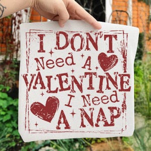 May include: A white square fabric with the text "I DON'T Need A VALENTINE I Need A NAP" in distressed red lettering. The design includes two heart shapes and star accents, perfect for Valentine's Day or a nap lover.