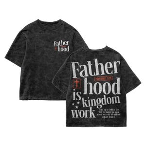 A flat lay showcasing both the front and back design of a dark charcoal vintage mineral wash t-shirt on a white background. The front features a small chest graphic that reads "Father hood" with a red cross icon. The back displays the full, large graphic reading "Father hood is kingdom work" in a distressed white font, accented by a red Proverbs 22:6 emblem, small stars, and the full biblical scripture quote.