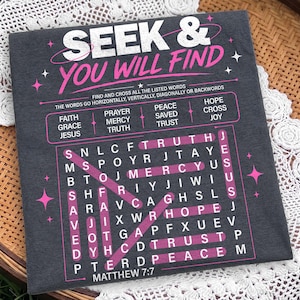 Close-up flat lay mockup of a dark charcoal shirt or fabric surface featuring a Christian “Seek & You Will Find” word search design in distressed white and pink. The artwork includes highlighted faith words, Matthew 7:7 scripture, star details, and a bold puzzle layout, displayed on a textured fabric background with cozy woven decor. Strong preview for digital shirt designs.