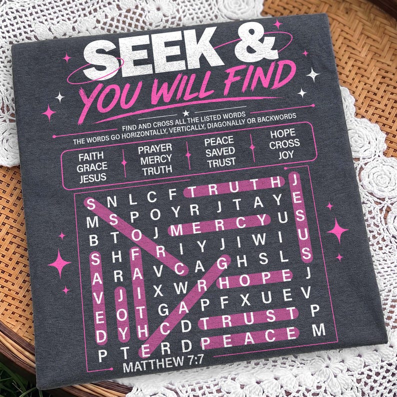 Close-up flat lay mockup of a dark charcoal shirt or fabric surface featuring a Christian “Seek & You Will Find” word search design in distressed white and pink. The artwork includes highlighted faith words, Matthew 7:7 scripture, star details, and a bold puzzle layout, displayed on a textured fabric background with cozy woven decor. Strong preview for digital shirt designs.
