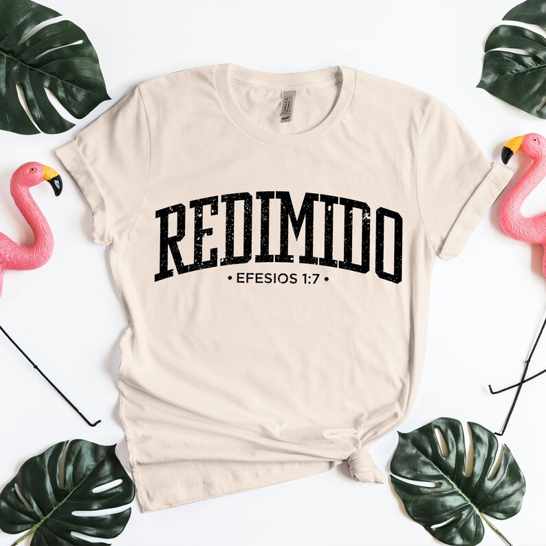 Neutral cream t-shirt mockup displaying the Spanish word “REDIMIDO” in arched distressed lettering with Efesios 1:7 printed beneath. The shirt is arranged in a flat lay style with a tied knot at the hem, framed by decorative green monstera leaves and pink flamingos on a white background. This mockup presents a soft, uplifting Christian message focused on hope, faith, and inspirational scripture-based design.