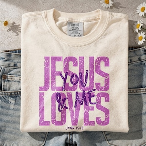 Folded cream Comfort Colors t-shirt displaying a distressed purple “Jesus Loves You & Me” Christian graphic with John 15:13 Bible verse. The mockup is styled on a textured neutral surface with scattered white daisy flowers and light denim jeans, creating a soft feminine aesthetic. This lifestyle scene highlights a faith-inspired graphic tee perfect for Christian apparel listings and inspirational religious shirt designs.