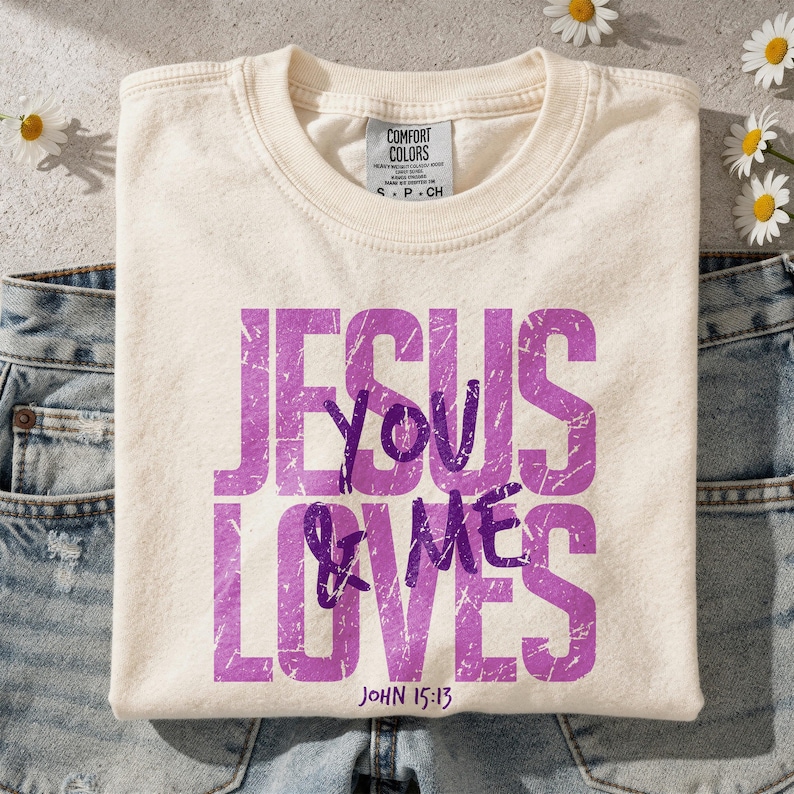 Folded cream Comfort Colors t-shirt displaying a distressed purple “Jesus Loves You & Me” Christian graphic with John 15:13 Bible verse. The mockup is styled on a textured neutral surface with scattered white daisy flowers and light denim jeans, creating a soft feminine aesthetic. This lifestyle scene highlights a faith-inspired graphic tee perfect for Christian apparel listings and inspirational religious shirt designs.