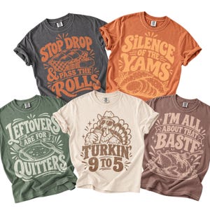 May include: Five short-sleeve crew neck t-shirts in charcoal, orange, sage green, cream, and brown. Each shirt features a unique graphic print with text: Stop Drop & Pass The Rolls, Silence Of The Yams, Leftovers Are For Quitters, Turkin' 9 To 5, and I'm All About That Baste.