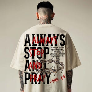 Back view of a model in an oversized cream t-shirt with neck tattoos, displayed against a neutral background. The shirt features a bold back print reading "Always Stop and Pray" layered over red text, accompanied by a crown of thorns graphic and Philippians 4:6 scripture text. Modern, edgy urban Christian streetwear tee for an impactful daily faith statement.