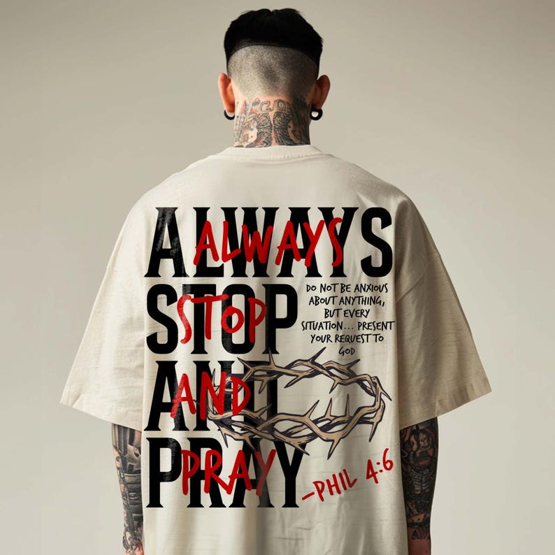 Back view of a model in an oversized cream t-shirt with neck tattoos, displayed against a neutral background. The shirt features a bold back print reading "Always Stop and Pray" layered over red text, accompanied by a crown of thorns graphic and Philippians 4:6 scripture text. Modern, edgy urban Christian streetwear tee for an impactful daily faith statement.