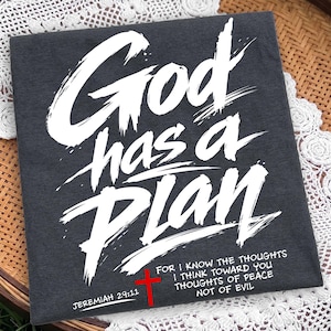 Dark folded apparel or canvas-style flat lay on a lace and woven rattan background, showcasing a high-contrast Christian streetwear design. The graphic reads “God has a Plan” in large distressed white brush lettering, with a red cross, Jeremiah 29:11 reference, and shortened scripture quote along the bottom for a bold faith-inspired printable mockup.