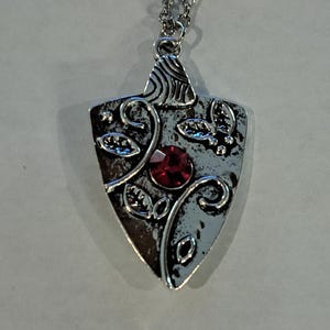 May include: Silver-toned pendant necklace with a shield-shaped pendant. The pendant features a red gemstone at the center, surrounded by leaf and swirl designs. The necklace has a delicate chain.