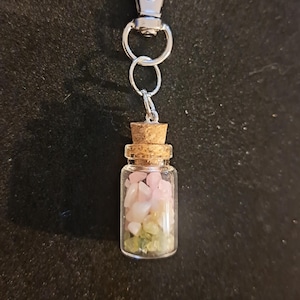 May include: A small glass bottle filled with pink and green gemstones, topped with a cork, and attached to a silver-colored keychain clip. The bottle is clear, allowing the colorful stones to be visible.