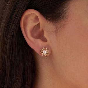 May include: A gold stud earring featuring a central round white pearl surrounded by a halo of small clear stones arranged in a starburst pattern. The earring is secured to the earlobe, showcasing a metallic gold setting and a lustrous pearl finish.