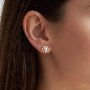 May include: A gold stud earring featuring a central round white pearl surrounded by a halo of small, clear, sparkling stones arranged in a circular pattern, secured to the earlobe with a gold post and backing mechanism.