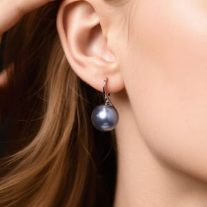 Könnte beinhalten: Ein einzelner silberner Ohrring mit einer großen, runden, grauen Perle. Der Ohrring ist eingehakt und hängt am Ohr. Die Perle hat eine glatte, reflektierende Oberfläche. Der Hintergrund ist unscharf, wodurch die Aufmerksamkeit auf den Schmuck gelenkt wird.