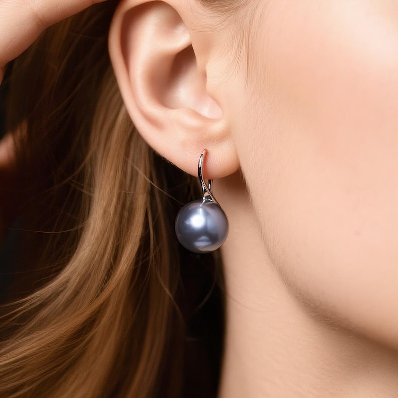 May include: A single silver earring with a large, round, gray pearl. The earring is hooked and hangs from the ear. The pearl has a smooth, reflective surface. The background is blurred, focusing attention on the jewelry.