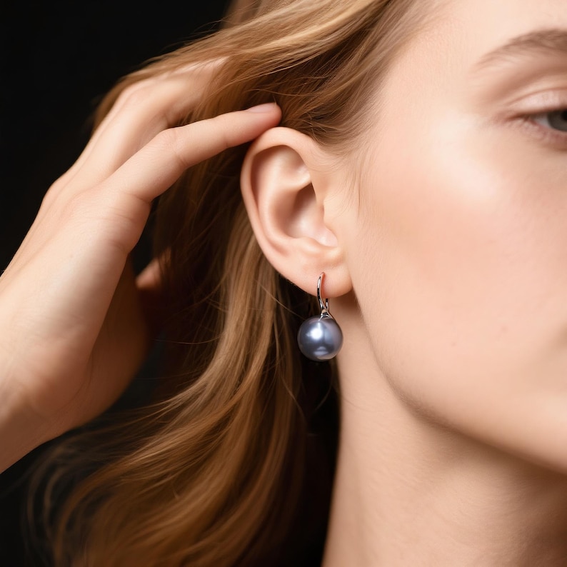 May include: A close-up of a silver earring with a large, dark gray pearl. The earring is hanging from the ear of a person with blonde hair. The background is black.
