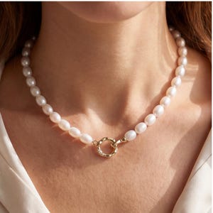May include: A necklace composed of oval-shaped white pearls strung together, featuring a gold-toned circular clasp at the front. The clasp has an irregular, textured surface and is accented with small, clear embedded stones.