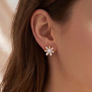 Puede incluir: Un pendiente de botón en forma de flor con pétalos blancos y una perla blanca central. El pendiente está sujeto al lóbulo de la oreja y presenta un marco de metal dorado alrededor de cada pétalo y una perla redonda y lisa en el centro.
