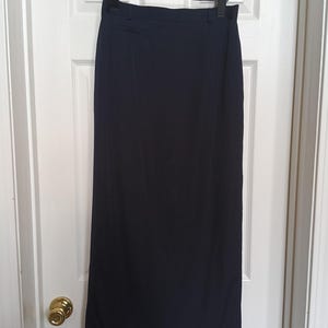 May include: A navy blue, floor-length skirt hangs on a wooden hanger. The skirt has a high waist and a straight silhouette. The fabric appears to be a smooth, lightweight material. The skirt is displayed against a white door.