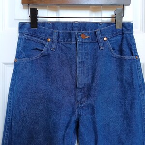 May include: Close-up of blue denim jeans hanging on a wooden hanger. The jeans have classic five-pocket styling with copper rivets and a button closure. The denim appears to be a medium wash, with visible stitching details.