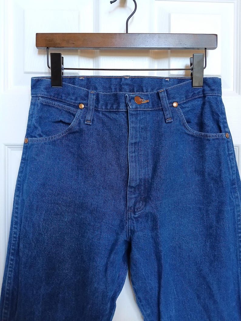 May include: Close-up of blue denim jeans hanging on a wooden hanger. The jeans have classic five-pocket styling with copper rivets and a button closure. The denim appears to be a medium wash, with visible stitching details.