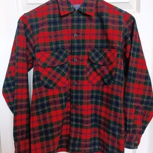 May include: A red, green, and navy plaid flannel shirt with two chest pockets and a collared neckline. The long-sleeved shirt is buttoned down the front and has buttoned cuffs. The shirt is hanging on a wooden hanger.