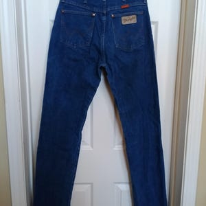 May include: Dark blue denim jeans with the Wrangler logo patch on the back pocket. The jeans have a classic five-pocket design with a straight leg. The jeans are hanging on a wooden hanger.