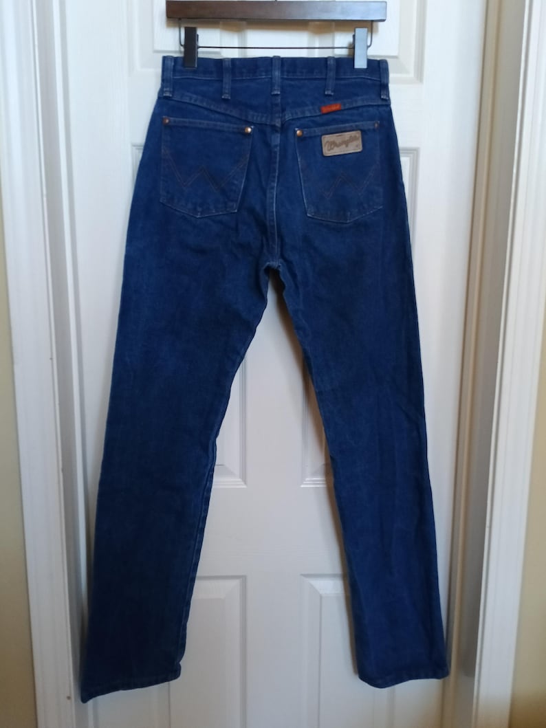 May include: Dark blue denim jeans with the Wrangler logo patch on the back pocket. The jeans have a classic five-pocket design with a straight leg. The jeans are hanging on a wooden hanger.