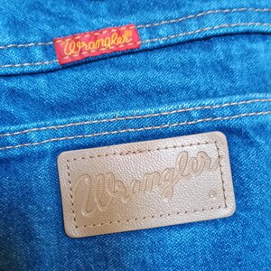 May include: Close-up of blue denim jeans with a red "Wrangler" label stitched on the back pocket. A brown leather patch with the "Wrangler" logo is also visible. The stitching is in a contrasting yellow thread.