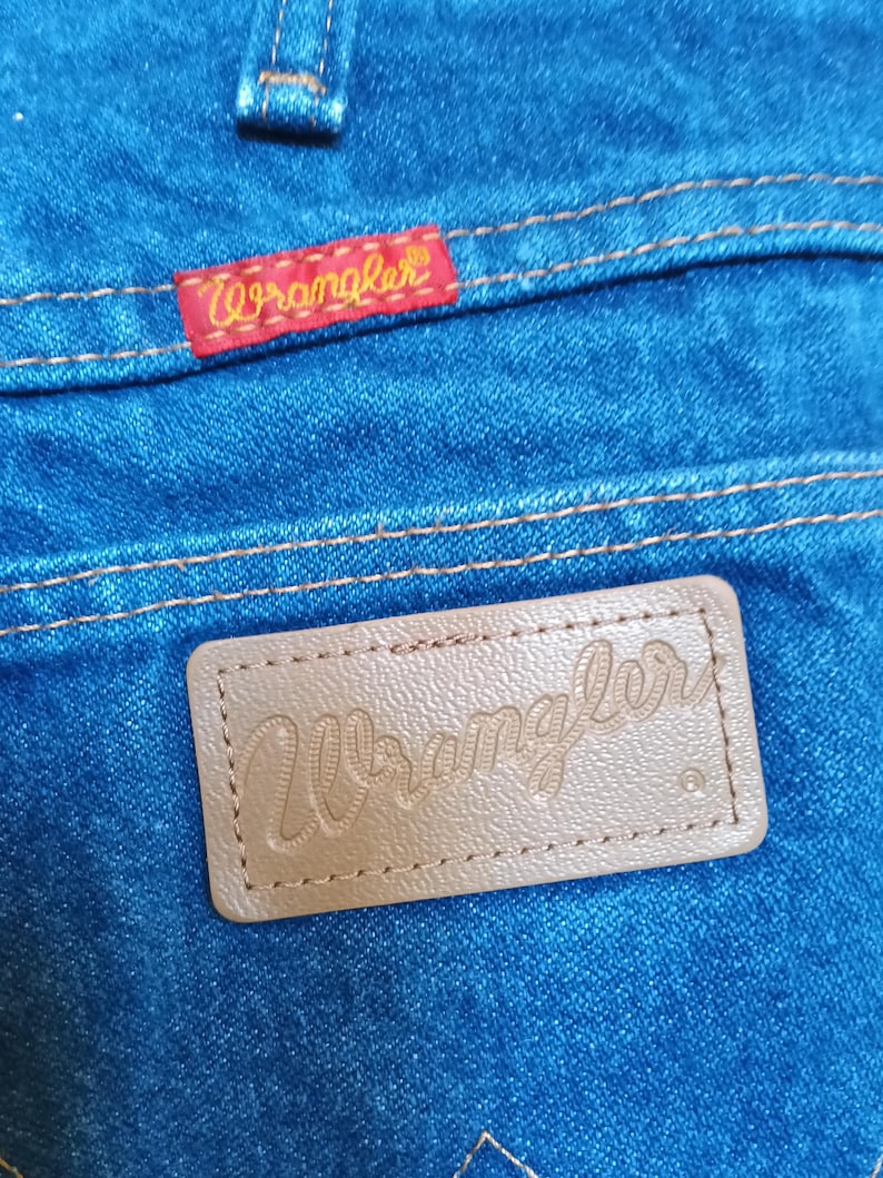 May include: Close-up of blue denim jeans with a red "Wrangler" label stitched on the back pocket. A brown leather patch with the "Wrangler" logo is also visible. The stitching is in a contrasting yellow thread.