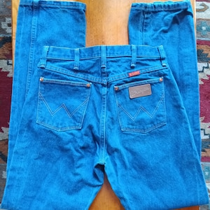 May include: Blue denim jeans with the Wrangler logo on the back pocket. The jeans have a classic five-pocket design, a zipper fly, and a straight leg. The back pockets feature the signature Wrangler 'W' stitching.