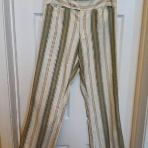May include: Cream-colored pants with vertical stripes in olive green and a thin gold stripe. The high-waisted pants have a wide waistband with two buttons. The pants are hanging on a wooden hanger.