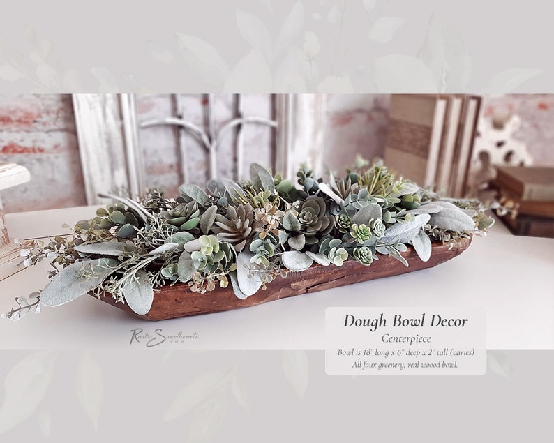 May include: A wooden dough bowl filled with faux greenery, including succulents and eucalyptus. The bowl is 18 inches long, 6 inches deep, and 2 inches tall. The text below the bowl reads 'Dough Bowl Decor Centerpiece Boul is 18' long x 6' deep x 2' tall (varies) All faux greenery, real wwood bowl.'