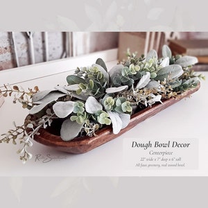 May include: A wooden dough bowl filled with faux greenery, including eucalyptus and lambs ear, for a farmhouse style centerpiece. The bowl is 22 inches wide, 7 inches deep, and 6 inches tall.  "Dough Bowl Decor Centerpiece" is written below the bowl.