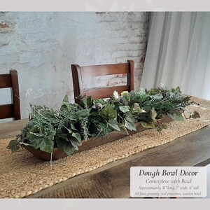 May include: A wooden dough bowl centerpiece filled with faux greenery and real pinecones. The centerpiece is approximately 31 inches long, 7 inches wide, and 6 inches tall. The bowl sits on a jute table runner, creating a rustic aesthetic. The text on the image reads "Dough Bowl Decor Centerpiece with Bowl".