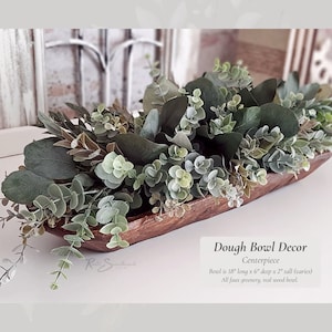 May include: A wooden dough bowl filled with faux greenery, including eucalyptus and other leafy plants. The bowl is 18 inches long, 6 inches deep, and 2 inches tall. "Dough Bowl Decor Centerpiece" and "Boul is 18" long x 6" deep x 2" tall (varies) All faux greenery, real wood bowl." are written below the bowl.