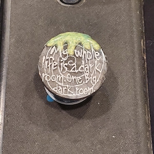 Puede incluir: Un soporte redondo y plateado para teléfono con el texto "My whole life is a dark room. One Big dark room." La parte superior tiene un diseño de pintura verde goteando. Está adherido a un teléfono negro.