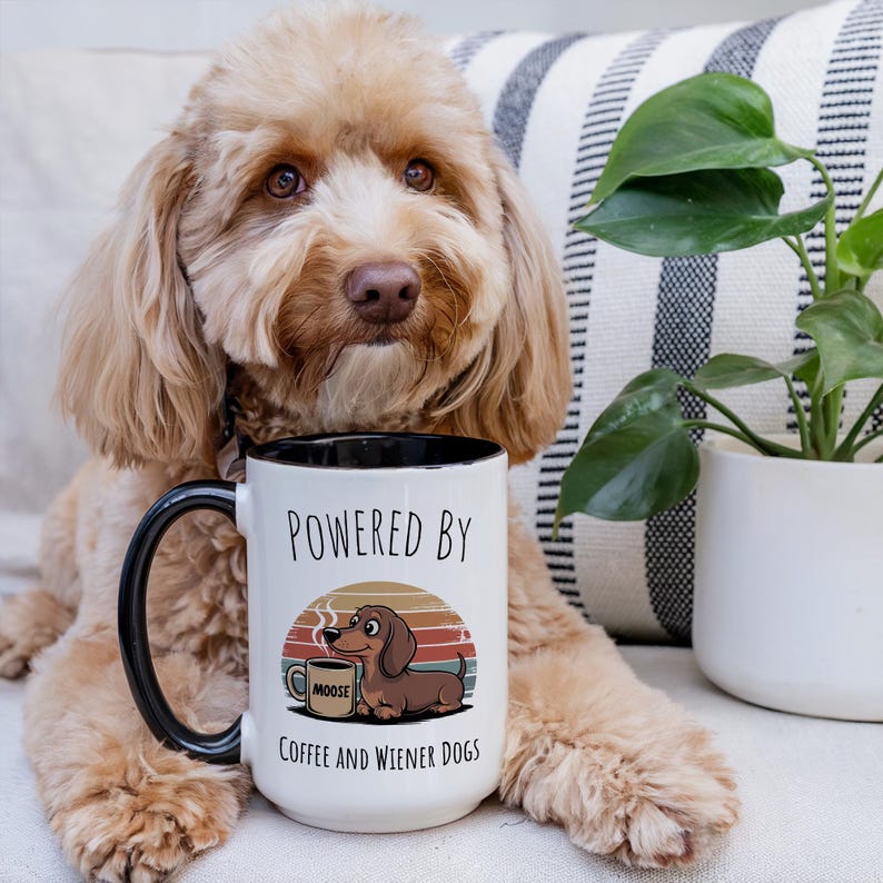 May include: White coffee mug with a black handle and rim, featuring the text "POWERED BY COFFEE AND WIENER DOGS." The mug has a cartoon illustration of a dachshund and a coffee cup. The mug is in front of a dog.