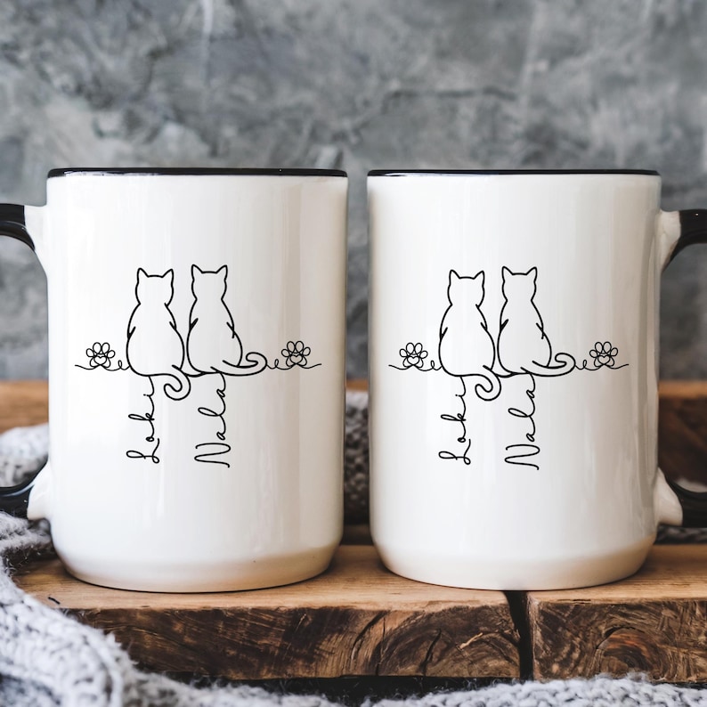 May include: Two white ceramic mugs with black rims and handles. Each mug features a black line drawing of two cats sitting side-by-side on a branch with flowers. The names "Leo" and "Nala" are written below the cats in a script font.