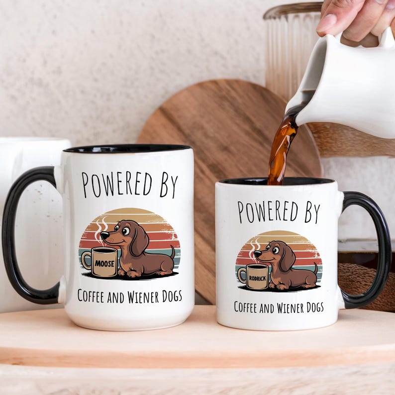 May include: Two white ceramic mugs with black rims and handles. The mugs feature a cartoon dachshund dog with a coffee cup and the text "POWERED BY COFFEE AND WIENER DOGS". One mug says "MOOSE" and the other says "RODRICK".