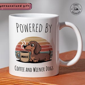 May include: White ceramic mug with a wraparound design featuring a cartoon dachshund dog, a coffee cup, and the text "POWERED BY COFFEE AND WIENER DOGS." The mug has a large handle and the word "MOOSE" on the coffee cup.