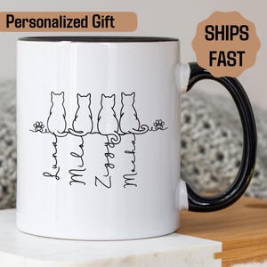 May include: White ceramic mug with a black handle and rim. The mug features a line of four cat silhouettes with names below them. The text "Personalized Gift" is at the top left. The mug is decorated with floral accents.