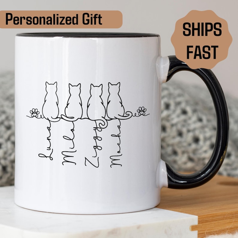 May include: White ceramic mug with a black handle and rim. The mug features a line of four cat silhouettes with names below them. The text "Personalized Gift" is at the top left. The mug is decorated with floral accents.