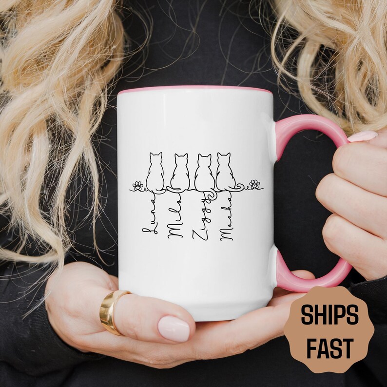 May include: White ceramic mug with a pink handle and interior. The mug features a black line drawing of five cats sitting in a row, with the names Luna, Mila, Ziggy, and Mocha below. The mug also has the text "SHIPS FAST".