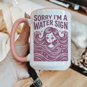 May include: White ceramic mug with a pink handle and rim. The mug features the text "SORRY I'M A WATER SIGN" in purple, along with a purple illustration of a woman with long flowing hair. The mug also has the astrological symbols for Cancer, Pisces, and Scorpio.