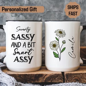 May include: Two white ceramic mugs with black handles and rims. One mug has the text "Seventy SASSY AND A BIT Smart ASSY" in black script. The other mug features a daisy floral design and the name "Camila".