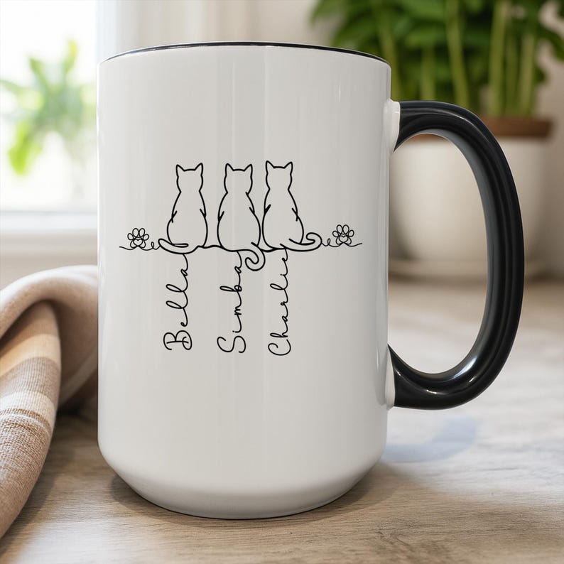 May include: White ceramic mug with a black handle and rim. The mug features a black line drawing of three cats sitting on a line, with the names "Bella", "Simba", and "Charlie" written below each cat.