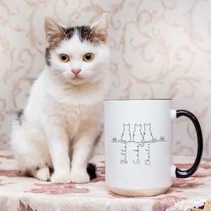 May include: A white ceramic mug with a black handle and rim, featuring a line drawing of three cats and the names "Bella", "Simba", and "Charlie". A white and gray cat sits beside the mug. The mug is on a floral patterned surface.