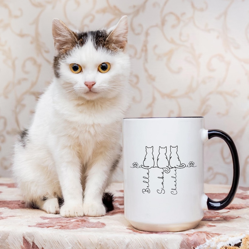 May include: A white ceramic mug with a black handle and rim, featuring a line drawing of three cats and the names "Bella", "Simba", and "Charlie". A white and gray cat sits beside the mug. The mug is on a floral patterned surface.