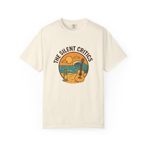May include: Cream-colored t-shirt featuring a circular graphic with the text "THE SILENT CRITICS." The graphic depicts a beach scene with a guitar, bottle, ocean waves, and sun. The shirt is made of a soft material.